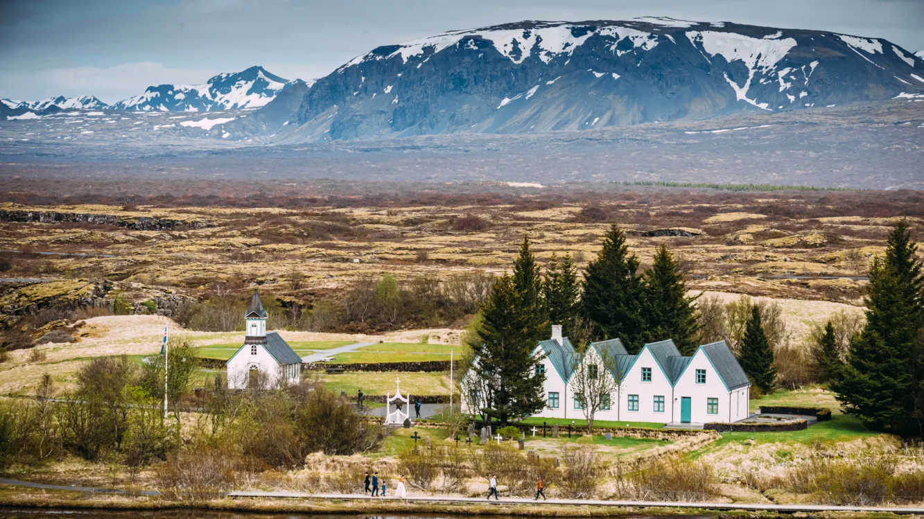 Thingvellir Kirche Island Thingvellir Kirche Island;
Kirche und 5 weiße Häuser mit grünem Dach, die aneinandergereiht sind
Im Hintergrund kahle und karge Ebene, dahinter Berge leicht mit Schnee bedeckt